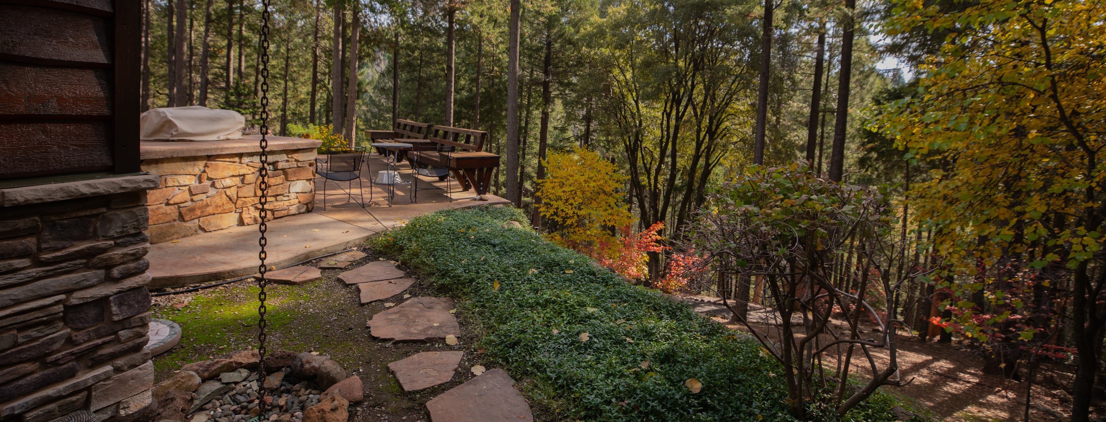 Stone patio and flagstone path beside a home, with low groundcover and fall-colored trees in a pine forest