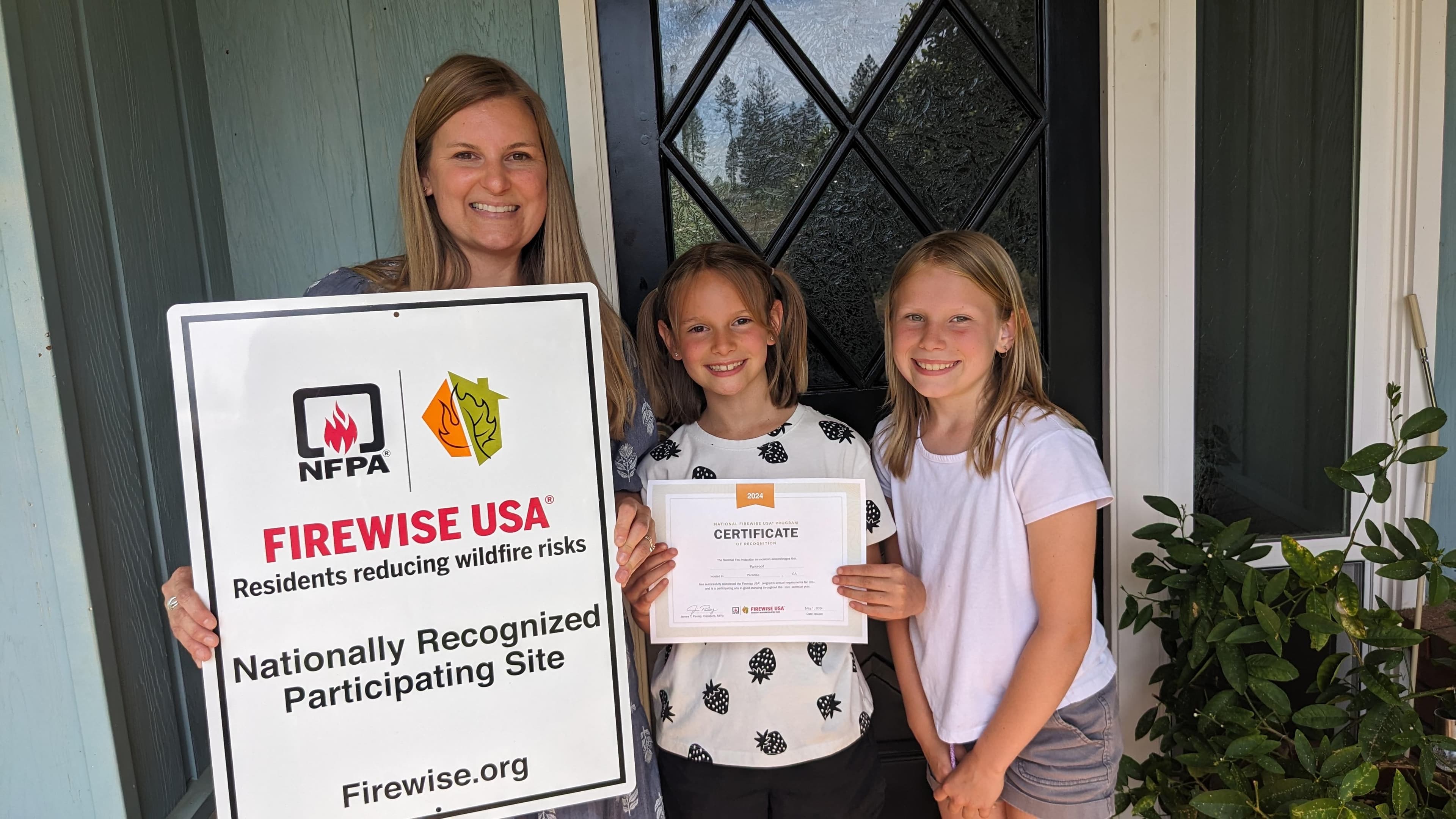 Family proudly displaying their Firewise USA Nationally Recognized Participating Site sign and 2024 certificate