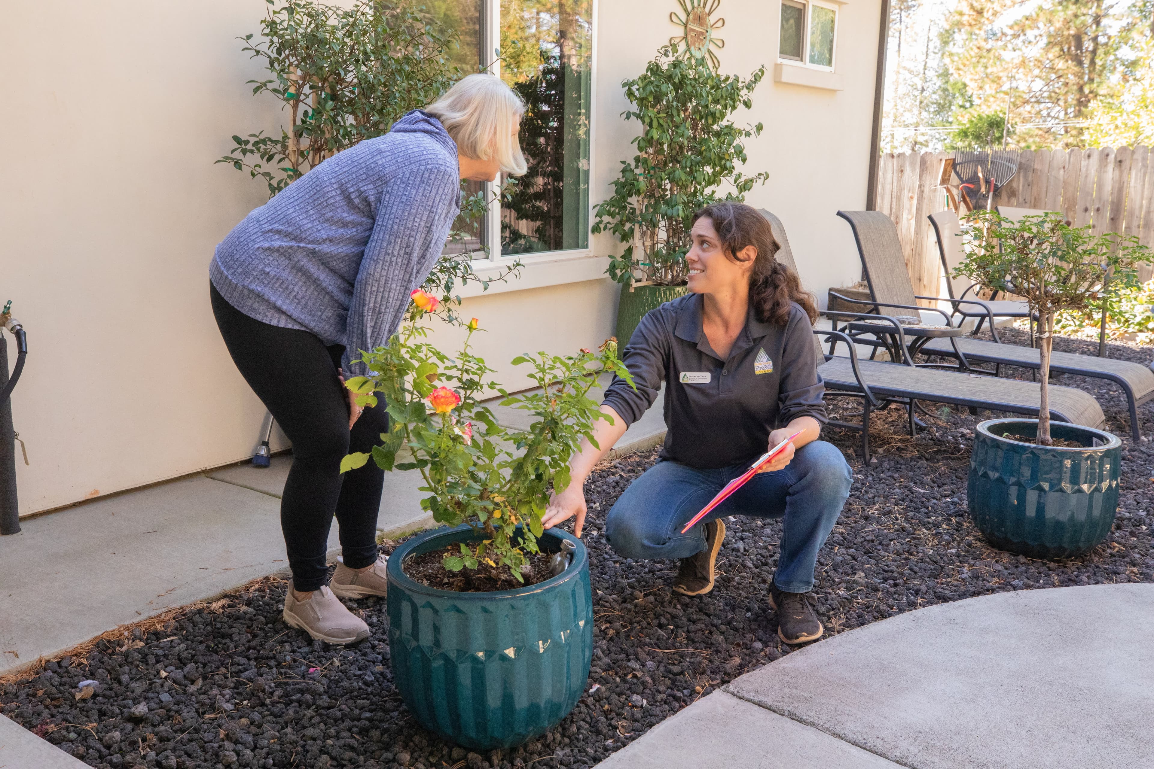 A BCFSC staff member with a clipboard crouches beside a potted rose, talking with a resident in their backyard